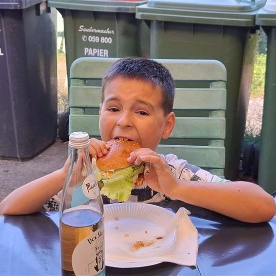 Ein junger Junge sitzt an einem Tisch und isst einen Hamburger, vor ihm steht eine Flasche Bier und ein Papierteller. Hinter ihm befinden sich grüne Mülleimer.