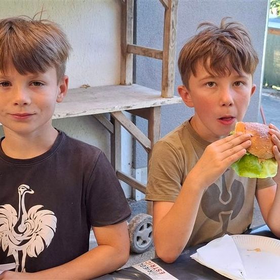 Zwei junge Jungen sitzen an einem Tisch, einer isst ein Sandwich und der andere hält eine Flasche.