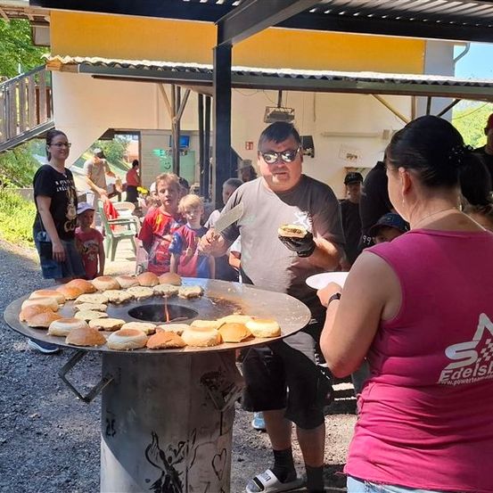 Ein Mann grillt Essen im Freien bei einer Veranstaltung, während eine Frau in einem rosa Shirt in der Nähe steht. Mehrere Menschen, einschließlich Kinder, sind um sie herum versammelt und beobachten.