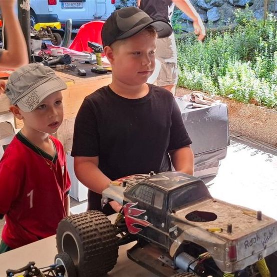 Zwei junge Jungen stehen neben einem ferngesteuerten Spielzeug-Truck, wobei einer einen Baseball-Cap trägt und der andere ein rotes Shirt. Dahinter steht eine Person in der Nähe eines Tisches mit verschiedenen Gegenständen darauf.