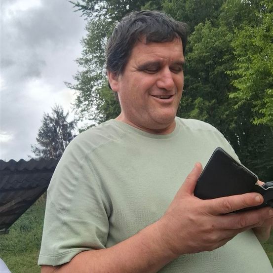 Ein Mann in einem hellgrünen T-Shirt lächelt und hält einen schwarzen Geldbeutel in den Händen. Er steht in einem Grasbereich mit Bäumen im Hintergrund.