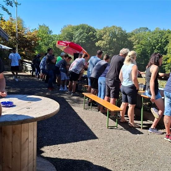 Eine Gruppe von Menschen versammelt sich um einen Holztisch und Bänke im Freien. Einige stehen, andere sitzen. Ein roter Regenschirm ist in der Nähe und ein Mann geht im Hintergrund.