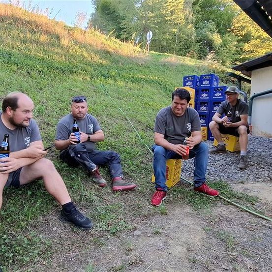 Vier Männer sitzen auf dem Boden und halten Bierflaschen in der Hand. Sie sind draußen, mit einem grasbewachsenen Hügel und einem Gebäude im Hintergrund.