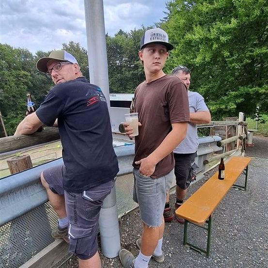 Drei Männer stehen im Freien, einer hält eine Flasche, ein anderer einen Becher und der dritte hat eine Flasche auf einer Bank. Sie sind in der Nähe eines Zauns und Bäumen.