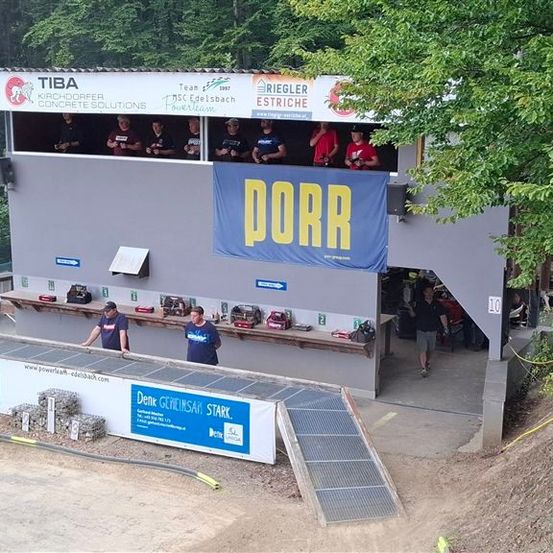 Eine Baustelle mit einem Banner mit der Aufschrift 'DORR'. Mehrere Personen sind im Innenraum des Standes, und andere arbeiten draußen. Die Baustelle ist von Bäumen umgeben und hat eine schräge Oberfläche.