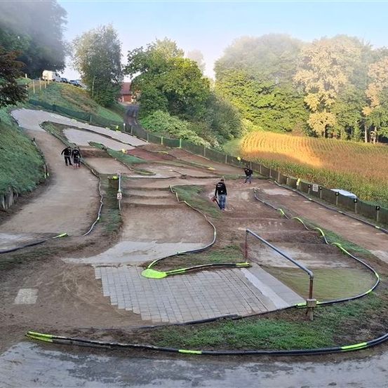 Eine Dirtbike-Strecke mit einigen Leuten, die darauf fahren. Die Strecke ist von Gras und Bäumen umgeben. Es gibt einige Stangen und Barrieren entlang der Strecke.