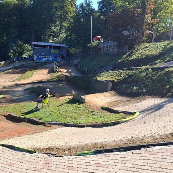 Eine Dirtbike-Strecke mit einer Person, die auf einem Stuhl in der Mitte der Strecke sitzt. Ein Gebäude mit dem Wort 'DORA' befindet sich auf der linken Seite.
