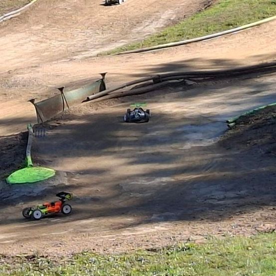 Zwei ferngesteuerte Autos rasen auf einer Erdrennstrecke mit einem grünen Objekt in der Nähe. Die Strecke hat eine Steigung und eine Barriere an der Seite.