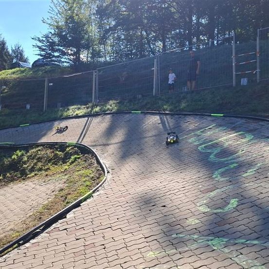 Eine kurvenreiche Ziegelstraße mit zwei ferngesteuerten Autos, umgeben von einem Metallzaun und Bäumen. Zwei Personen stehen in der Nähe des Zauns und beobachten die Autos.