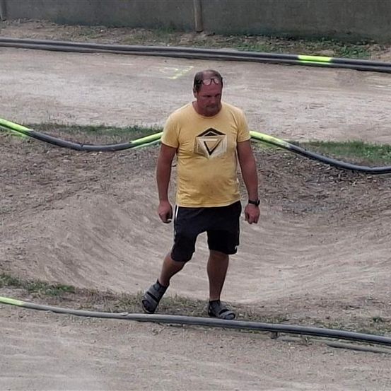 Ein Mann in einem gelben T-Shirt und schwarzen Shorts läuft auf einer Erdstrecke mit einer Kurve und gelben Markierungen. Er ist kahl und trägt eine Brille.