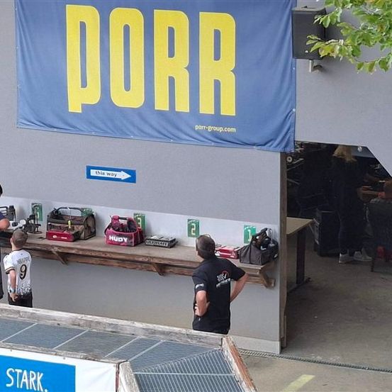 Eine Gruppe von Menschen steht vor einem Banner mit dem Wort PORR. Das Banner hängt an der Wand. Auf dem Tisch befinden sich mehrere Gegenstände, darunter ein Werkzeugkasten, eine rote Box und eine schwarze Tasche. Außerdem sitzen mehrere Personen auf Stühlen.