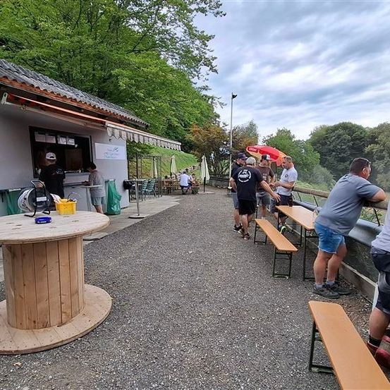 Eine Gruppe von Menschen versammelt sich in einem Restaurant im Freien mit Blick auf den Wald. Einige sitzen auf Bänken, andere stehen. Eine Person steht vor einer Theke.