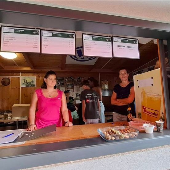 Drei Frauen stehen hinter einer Theke in einem kleinen Laden. Eine Frau trägt ein rosa Shirt und hält einen Ordner. Eine andere trägt ein schwarzes Shirt und hat die Arme verschränkt. Eine dritte trägt ein graues Shirt. Speisekarten hängen über ihnen.
