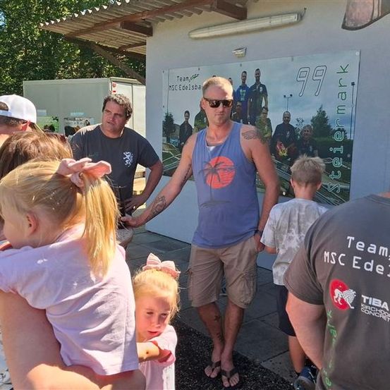 Eine Gruppe von Menschen, einschließlich Kinder, steht vor einem Gebäude mit einem Plakat. Der Mann in der Mitte trägt eine Sonnenbrille und ein ärmelloses Shirt.