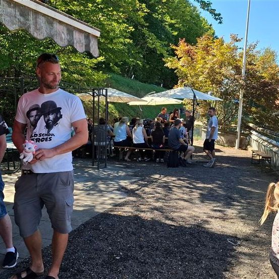 Eine Gruppe von Menschen versammelt sich in einem Außenbereich, möglicherweise einem Park, mit einem Mann, der ein Kuscheltier hält. Ein Mädchen geht auf der rechten Seite.