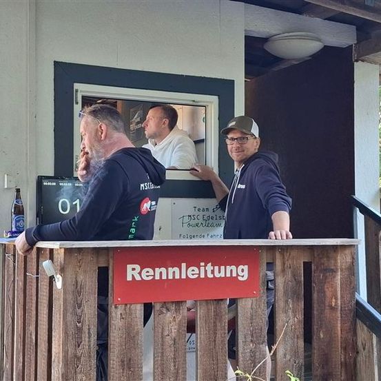 Drei Männer stehen auf einem hölzernen Balkon, einer von ihnen raucht. Ein Schild auf dem Balkon sagt Rennleitung. Ein Mann ist im Gebäude und schaut aus dem Fenster.