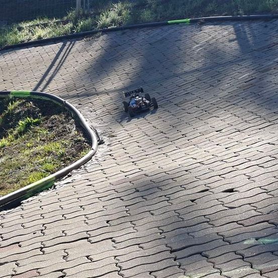 Ein ferngesteuertes Auto fährt auf einer gebogenen Ziegelstraße, mit einem Grasbereich links und einem schwarzen Randstein rechts.