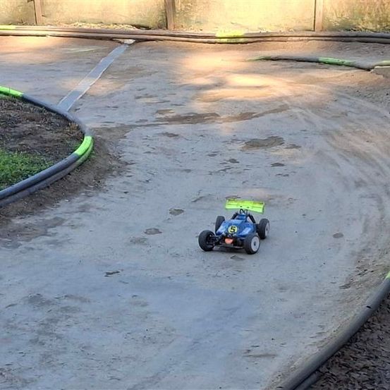 Ein blaues und grünes Spielzeugauto fährt auf einer unbefestigten Rennstrecke mit einer gelben Linie. Die Strecke ist von einem Grasbereich umgeben.