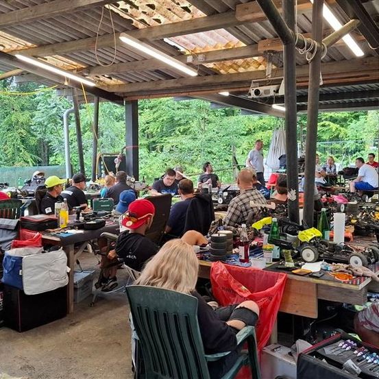 Eine Gruppe von Menschen versammelt sich in einer Werkstatt, wo sie an verschiedenen Projekten arbeiten. Die Werkstatt ist mit Werkzeugen, Geräten und Materialien gefüllt. Einige Leute sitzen auf Stühlen, während andere stehen und an Tischen arbeiten. Die Werkstatt ist gut beleuchtet, und draußen sind Bäume zu sehen.