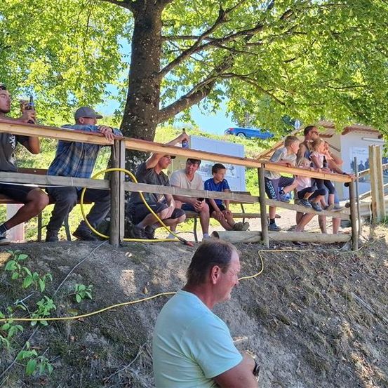 Eine Gruppe von Menschen sitzt auf einer Holzbank und beobachtet etwas. Sie tragen Freizeitkleidung und einige halten Flaschen. Die Bank befindet sich auf einem Hang mit einem gelben Kabel am Boden. Ein Mann sitzt vor ihnen auf dem Boden. Im Hintergrund befinden sich Bäume und ein Gebäude.
