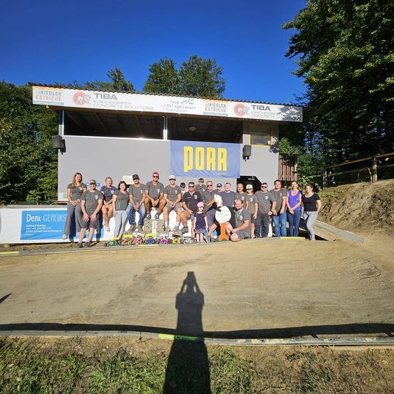 Eine Gruppe von Menschen posiert für ein Foto vor einem Gebäude mit einem Banner, auf dem 'POOR' steht. Sie tragen Freizeitkleidung und einige tragen Hüte. Hinter ihnen sind Bäume und ein Zaun.