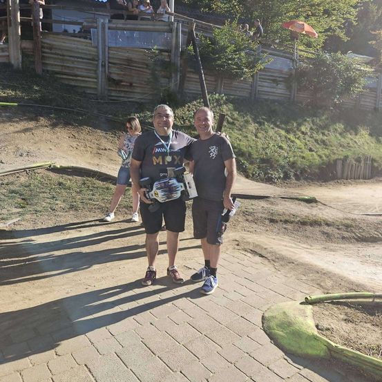 Zwei Männer stehen auf einem Ziegelweg, einer hält eine Kamera und der andere einen Pokal. Hinter ihnen geht eine Frau an einem Holzzaun vorbei. Bäume und ein Gebäude sind im Hintergrund zu sehen.