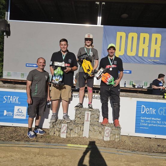 Vier Männer stehen auf einem Podium mit den Nummern 1, 2 und 3. Sie halten ferngesteuerte Autos und tragen Medaillen. Dahinter ist ein Banner mit dem Wort 'DORR' und ein Tisch mit einer Flasche.