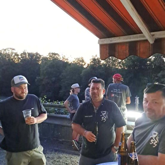 Vier Männer stehen unter einem Vordach, zwei halten Getränke, einer mit einer Bierflasche. Bäume und ein Zaun sind im Hintergrund.