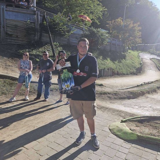 Ein Mann steht auf einem Ziegelweg, hält einen Pokal und ein grünes Objekt in der Hand, mit drei Personen hinter ihm und einer Erdbahn im Hintergrund.