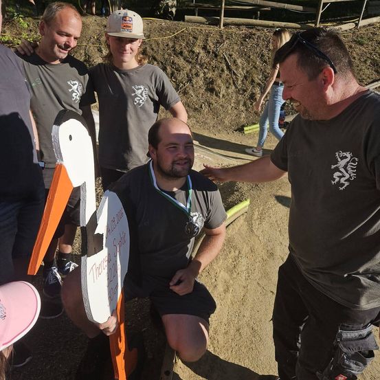 Eine Gruppe von Menschen steht auf einem Erdweg, einer von ihnen hält einen hölzernen Storch. Der Mann in der Mitte trägt eine Medaille und lächelt. Hinter ihnen geht eine Frau auf dem Weg.