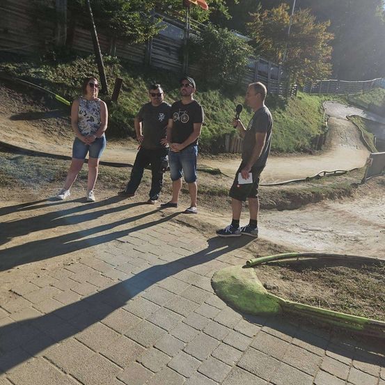 Vier Personen stehen auf einem Ziegelweg, einer hält ein Mikrofon. Sie sind in der Nähe einer Erdstrecke mit einem Holzzaun. Die Sonne scheint hell.