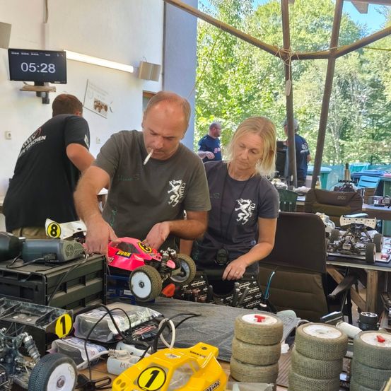 Ein Mann und eine Frau arbeiten in einer Werkstatt an einem pinkfarbenen Spielzeugauto. Der Mann raucht eine Zigarette, während die Frau ihn ansieht. Im Hintergrund sind andere Leute zu sehen, und auf dem Tisch liegen andere Spielzeuge und Werkzeuge.