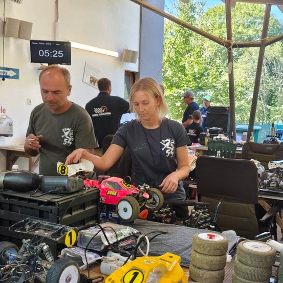 Ein Mann und eine Frau arbeiten an einem Tisch voller RC-Autos, wobei ein Auto die Nummer 8 und ein anderes die Nummer 1 hat. Sie sind von anderen Leuten und Ausrüstung umgeben.