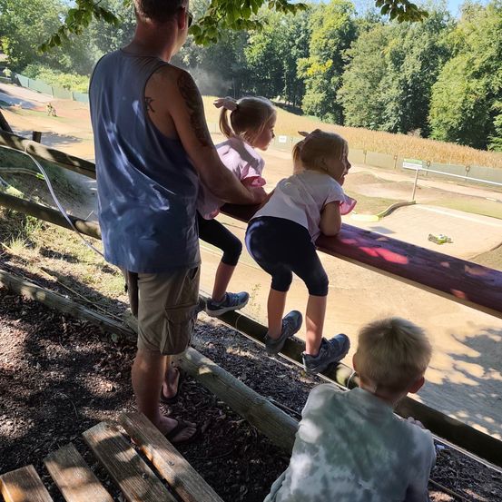 Ein Mann und drei Kinder stehen auf einem Holzzaun und beobachten eine Erdstrecke mit einem kleinen Auto darauf. Der Mann trägt ein ärmelloses Hemd und Shorts, während die Kinder Turnschuhe tragen.