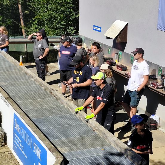 Eine Gruppe von Menschen versammelt sich um eine große Metallgitter, möglicherweise an einer Baustelle. Einige tragen Handschuhe und Mützen, einer hält ein Werkzeug. Links ist ein blauer Banner mit Text zu sehen.