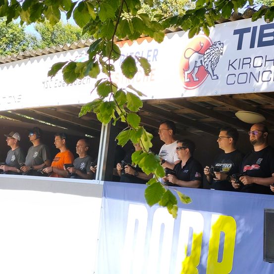 Eine Gruppe von Männern mit Sonnenbrillen und Waffen steht in einem Stand mit einem Schild, auf dem 'TIBI KIRCH CONCE' steht.