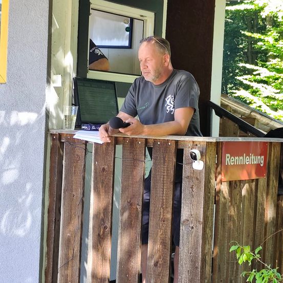Ein Mann mit einem Mikrofon steht auf einem Balkon, hält ein Stück Papier in der Hand und schaut nach unten. Hinter ihm befindet sich ein Monitor und ein Glas. Der Balkon hat ein hölzerne Geländer und ein Schild mit der Aufschrift 'Rennleutung'. Im Hintergrund ist ein Baum und ein Gebäude zu sehen.