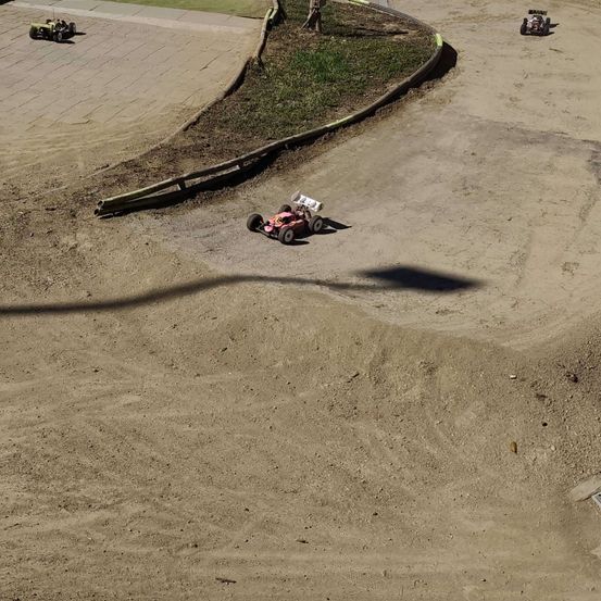 Eine Draufsicht auf eine Erdrennstrecke mit zwei Spielzeugautos im Rennen. Ein Auto liegt in Führung, während das andere hinterher ist. Die Strecke ist von Gras und einer Betonbarriere umgeben.