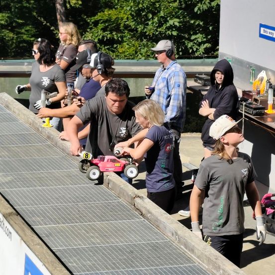 Eine Gruppe von Menschen versammelt sich um eine Rennstrecke und beobachtet ein pinkes Spielzeugautorennen. Die Strecke besteht aus Metall und hat eine Steigung. Die Teilnehmer tragen Handschuhe und einige haben Kopfhörer. Bäume und ein Gebäude sind im Hintergrund.
