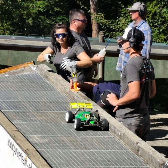 Eine Gruppe von Menschen beobachtet ein Rennen mit ferngesteuerten Autos auf einer Rennstrecke. Eine Frau hält eine Flasche und ein Mann trägt Kopfhörer.
