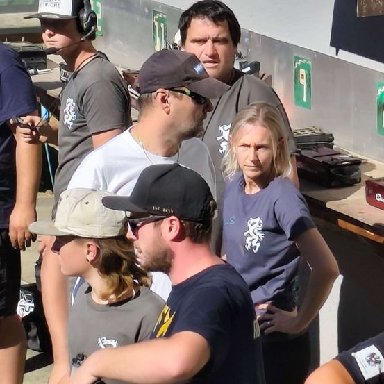 Eine Gruppe von Menschen in Hüten und Sonnenbrillen steht in einem Raum. Ein Mann mit schwarzem Hut schaut eine Frau mit ernster Miene an. Hinter ihnen steht ein Mann mit Kopfhörern und Mikrofon.