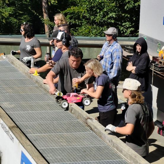 Eine Gruppe von Menschen versammelt sich um eine Rennstrecke und beobachtet, wie ein kleines rosa Spielzeugauto eine Steigung hinunterfährt. Einige tragen Handschuhe und Kopfhörer.