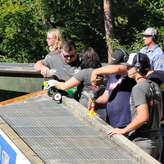 Eine Gruppe von Menschen versammelt sich um eine Rampe mit einem Metallgitter, möglicherweise zur Vorbereitung eines Rennens. Eine Person hält eine Sprühflasche, während eine andere ein Werkzeug in der Hand hat. Sie tragen alle Kopfhörer und scheinen sich auf die Aufgabe zu konzentrieren.