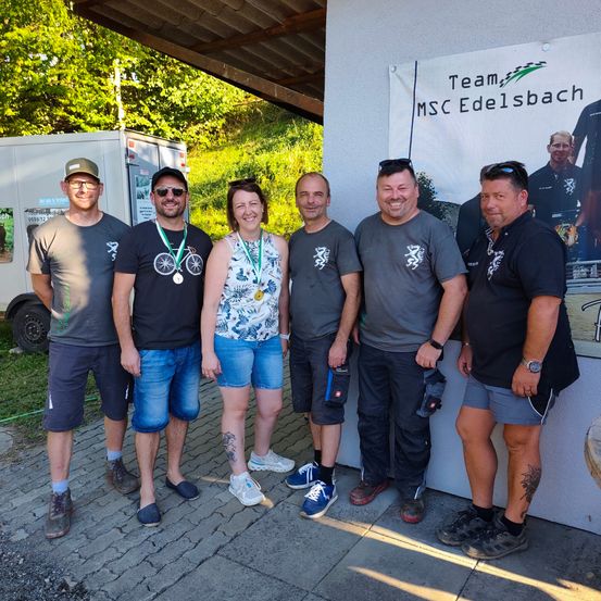 Eine Gruppe von sechs Personen, möglicherweise ein Radsportteam, steht vor einem Banner mit dem Text 'Team MSC Edelsbach'. Sie alle lächeln und scheinen für ein Foto zu posieren.