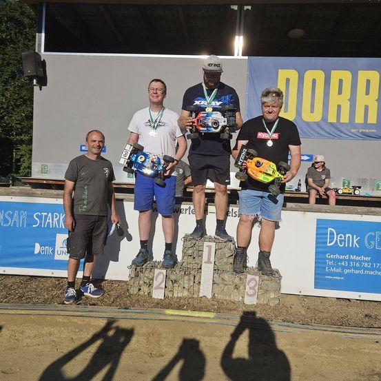 Vier Männer stehen auf einem Podium mit ihren ferngesteuerten Autos und tragen Medaillen. Das Podium ist mit 1, 2 und 3 nummeriert. Sie stehen auf einer Erdbahn mit einem Banner hinter ihnen.