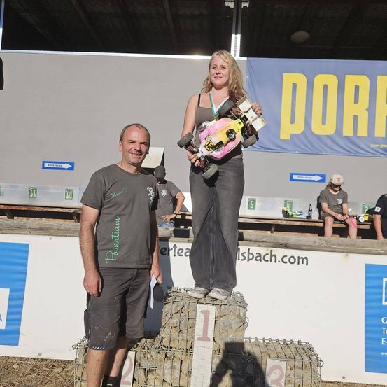 Eine Frau steht auf einem Haufen Steine und hält ein ferngesteuertes Auto, während ein Mann neben ihr steht. Sie stehen auf einem Podium bei einer Outdoor-Veranstaltung.