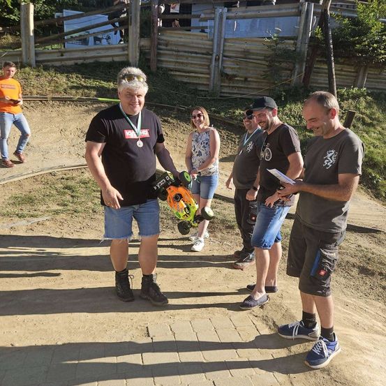Eine Gruppe von Menschen steht auf einer Erdbahn, ein Mann hält ein Spielzeugauto und eine Medaille, während andere zusehen. Hinter ihnen ist ein Holzzaun und ein Grasbereich sichtbar.