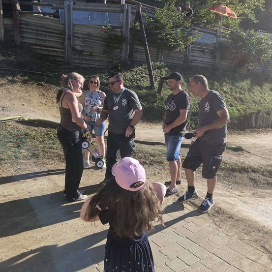 Eine Gruppe von Menschen steht auf einem Erdweg, eine Frau hält die Hand eines Kindes. Hinter ihnen ist ein Holzzaun und ein Grasbereich zu sehen. Zwei Männer tragen Mützen und einer Sonnenbrille. Eine Frau hält einen kleinen Wagen.