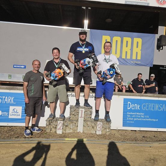 Vier Männer stehen auf einem Podium mit ferngesteuerten Autos und lächeln für ein Foto. Sie befinden sich bei einer Veranstaltung mit Bannern und Zuschauern im Hintergrund.