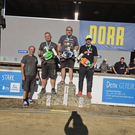 Vier Männer stehen auf einem Podium mit Trophäen, einer hält ein ferngesteuertes Auto. Sie lächeln und posieren für ein Foto. Dahinter steht auf einem Banner 'DORR'.
